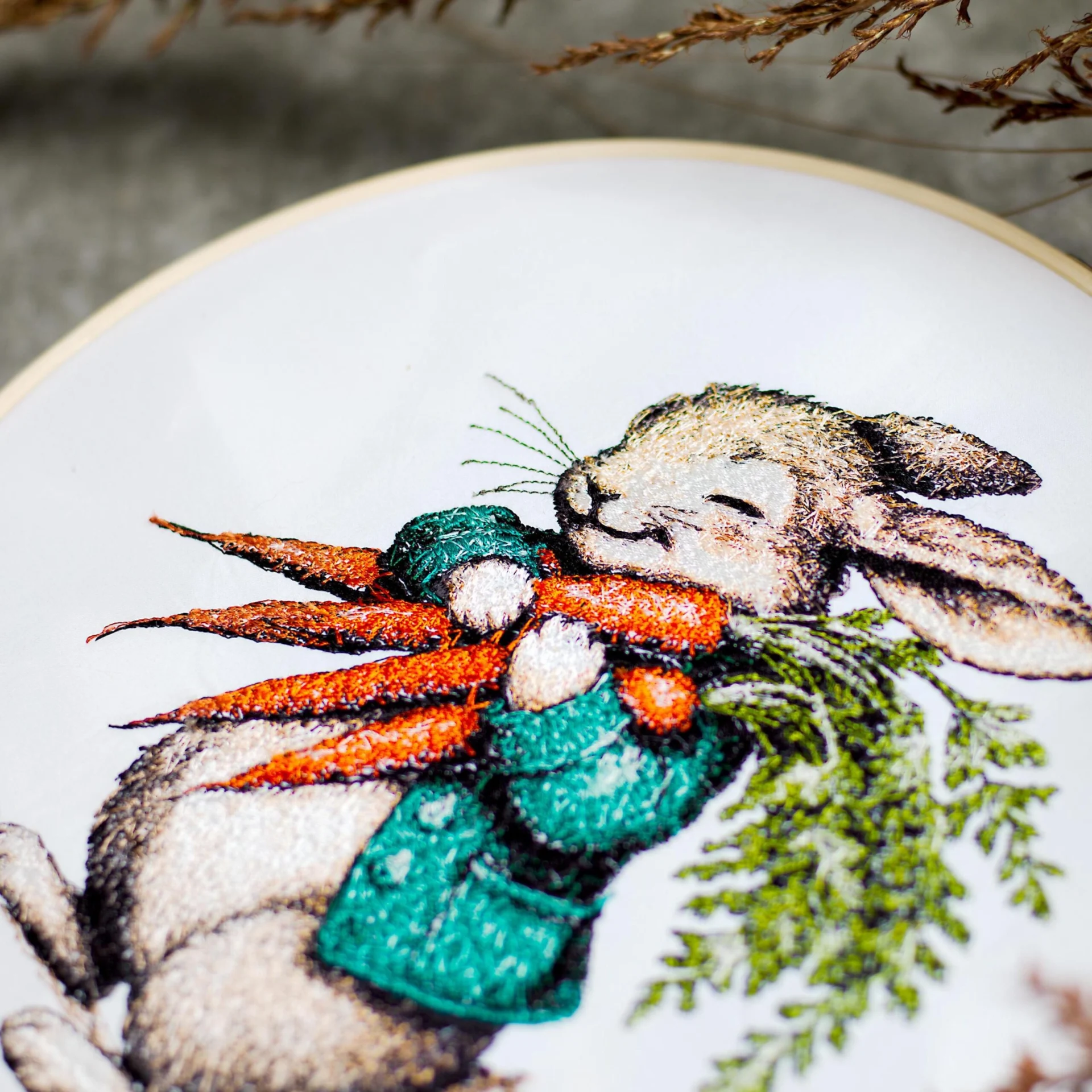 Close-up of a machine embroidery design featuring a joyful bunny in a textured green coat, cuddling a bundle of vibrant orange carrots with lush green tops. The intricate stitching captures fine fur details, expressive whiskers, and delicate shading, evoking a vintage storybook illustration. The rich thread textures and lifelike depth bring this whimsical woodland scene to life, perfect for Easter or heirloom embroidery projects.